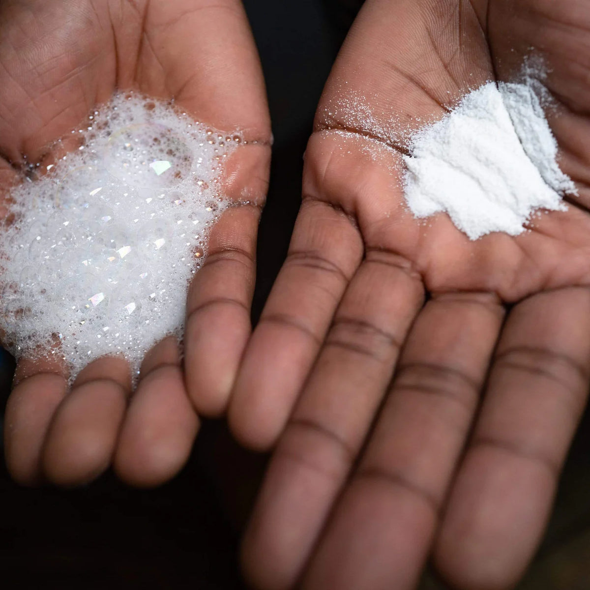 Two hands holding piles of white powder and white crystalline granules against a dark background.