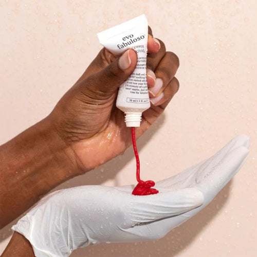 Hand holding a tube of evo fabuloso hair product with red substance dripping onto a white glove against a beige background.