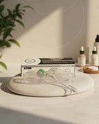 A derma roller set including a clear plastic derma roller, a green and white tube, and its black-and-white packaging box, styled on a round stone tray with spa bottles and a plant in the background.