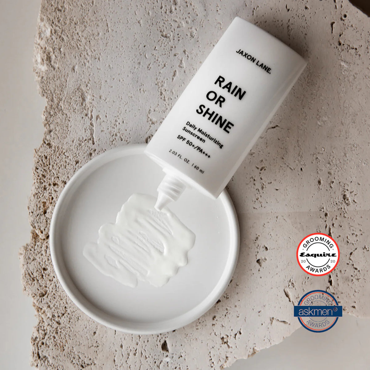A white squeeze tube of JASON LANE Rain or Shine daily moisturizing sunscreen lies diagonally on a textured stone surface above a shallow white dish containing white cream. Two certification badges are visible on the right side.